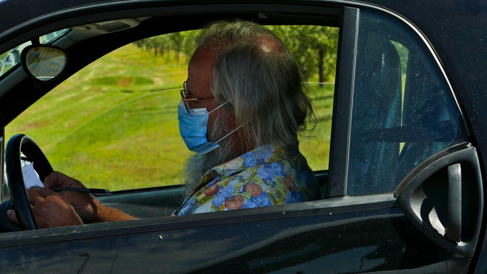 Un homme aux cheveux longs porte un couvre-visage et se trouve au volant de sa voiture. C'est l'été.