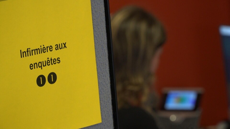 Gros plan sur une feuille de papier jaune sur laquelle sont écrits les mots « infirmière aux enquêtes ». La feuille est affichée sur une cloison délimitant un espace de travail. On aperçoit, à l’arrière-plan, une femme parlant au téléphone.