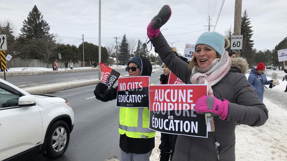 Des employés le long d'une rue saluent au passage des véhicules et tiennent des pancartes revendiquant « Aucune coupure en éducation ».