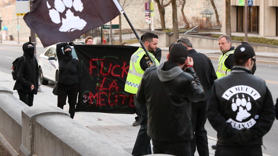 Des policiers s’interposent entre des membres de La Meute et trois contre-manifestants cagoulés. Ces derniers sont vêtus de noir et portent une banderole sur laquelle il est écrit : «Fuck La Meute».
