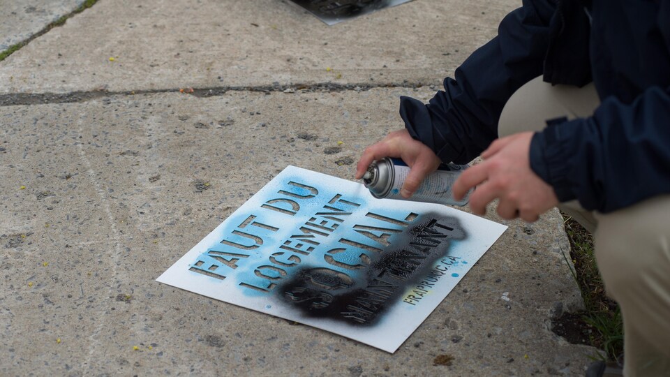 Un manifestant utilise de la peinture en aérosol et un pochoir pour tracer le message « Faut du logement social maintenant ».