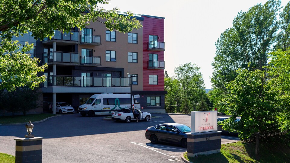 L'extérieur de l'établissement pour personnes aînées, le Manoir Liverpool, en été. On y voit des voitures stationnées et des personnes qui en sortent.