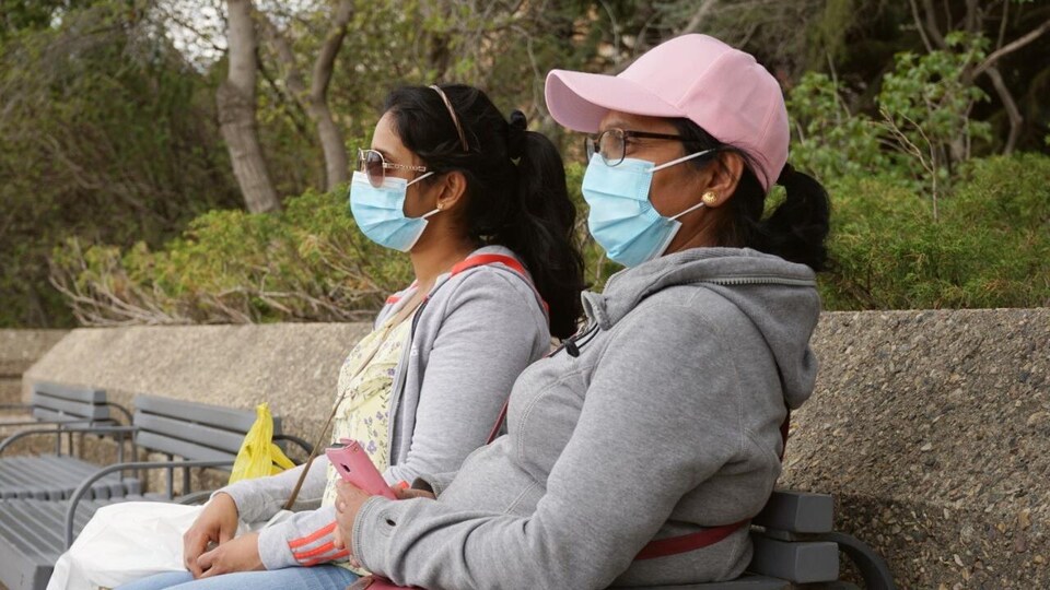 Deux femmes asises sur un banc à l'extérieur, avec des masques sur le visage.