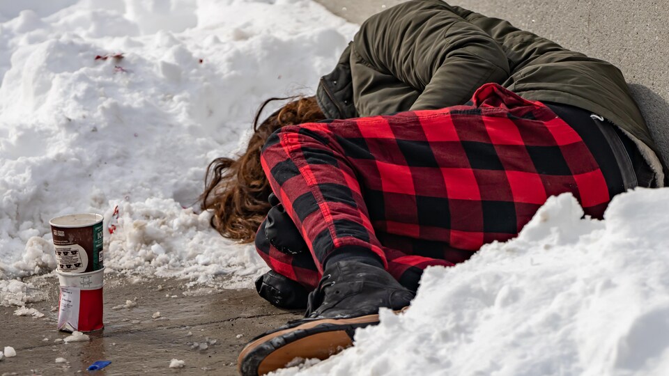 Un itinérant couché sur le sol, entouré de neige, à Ottawa. Près de lui, un vieux gobelet à café à usage unique.