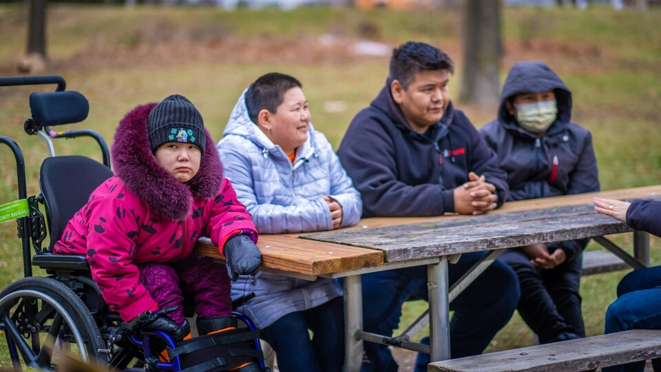 Une adolescente dans un fauteuil roulant et des membres de sa famille assis à ses côtés sur un banc de bois dans un parc.
