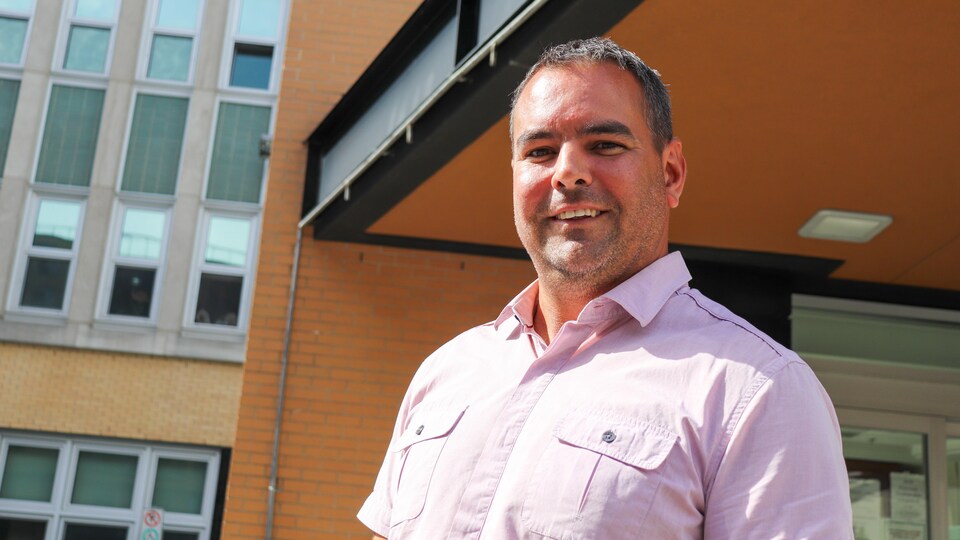 Portrait en contre-plongée d'Éric Lavoie. Il sourit et se tient debout devant un établissement de santé.