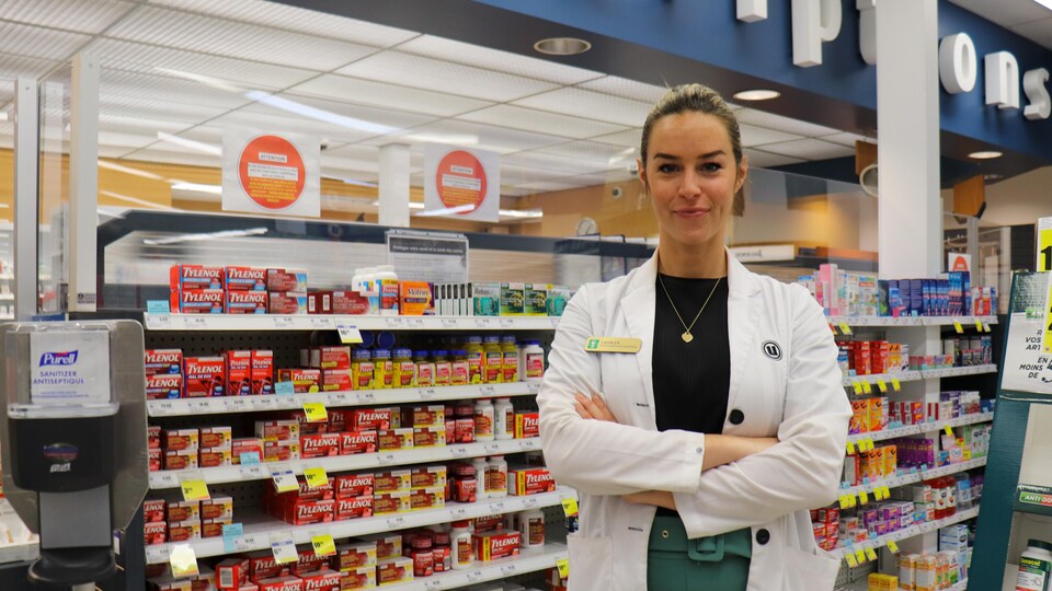 Camille Dupuis-Brousseau, vêtue d’un sarrau, se tient debout, les bras croisés, devant le comptoir des prescriptions d’une pharmacie. 