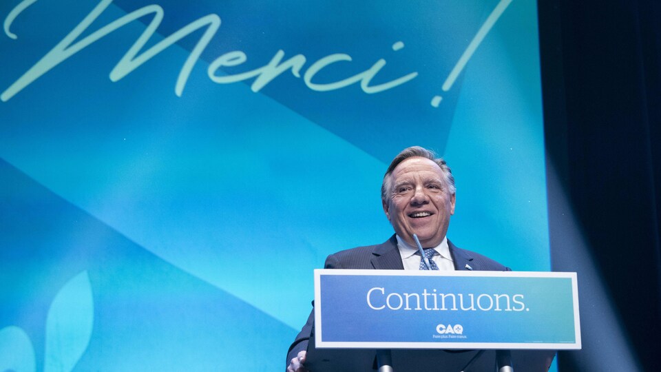 François Legault est debout derrière un lutrin où apparaît le mot « Continuons » et devant un grand écran à l'arrière-plan où apparaît le mot « Merci! ».