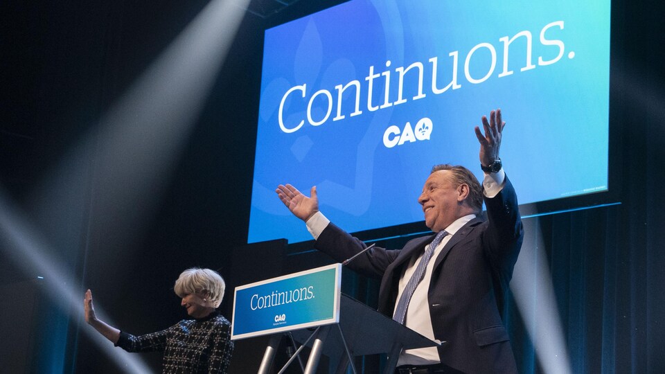 François Legault lève les bras en signe de victoire, derrière un lutrin, tandis qu'on voit son épouse Isabelle Brais faire un salut de la main.