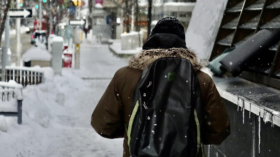Un piéton, de dos, marchant sur un trottoir légèrement enneigé.