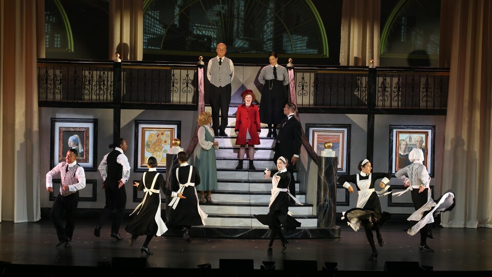 Des comédiens dansent et chantent sur la scène. Un grand escalier est placé au centre.