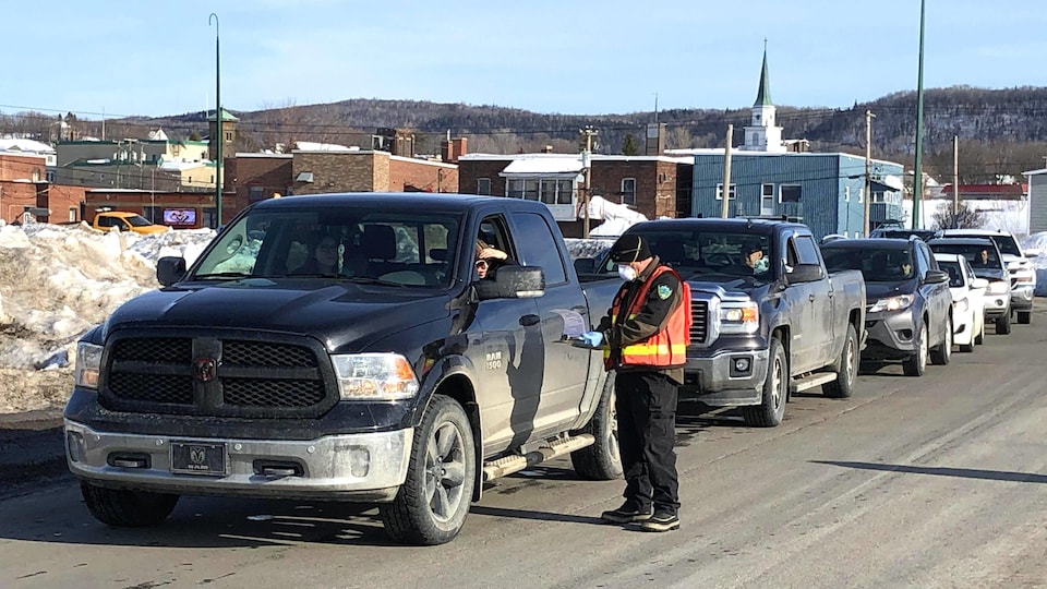 Un homme portant une veste orange et un masque respiratoire s'approche d'une automobile avec des documents.