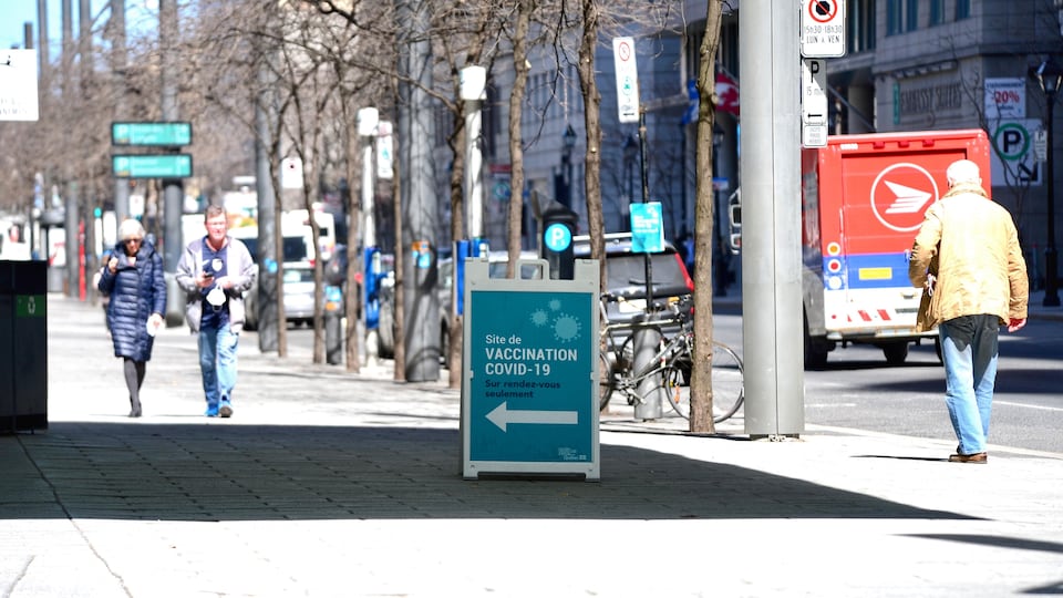 Des passants dans la rue marchent sous le soleil et une pancarte annonce une clinique de vaccination.