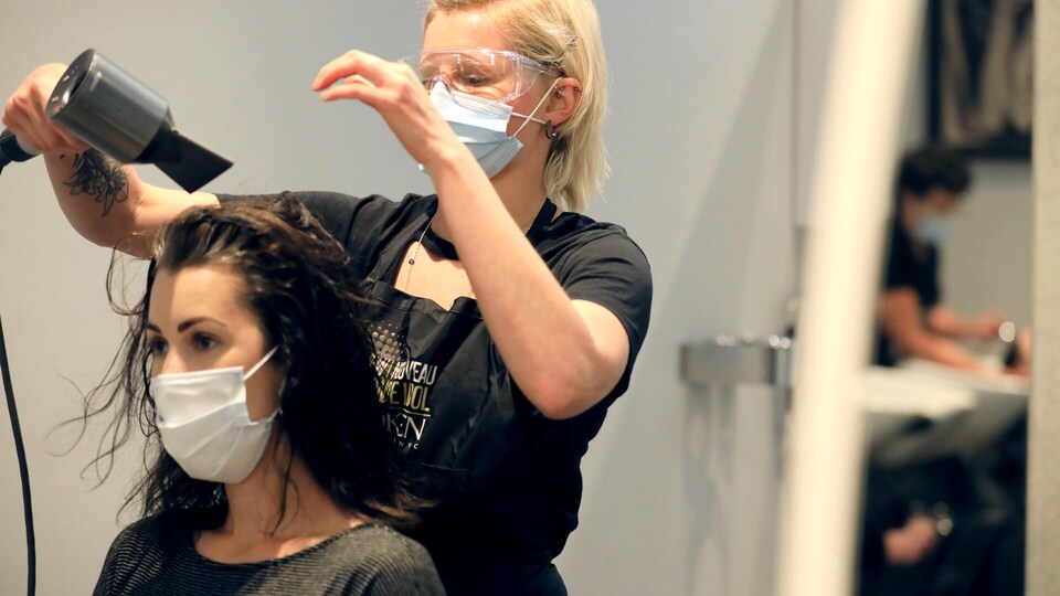 Une coiffeuse sèche les cheveux d'une cliente. Les deux femmes portent un masque.