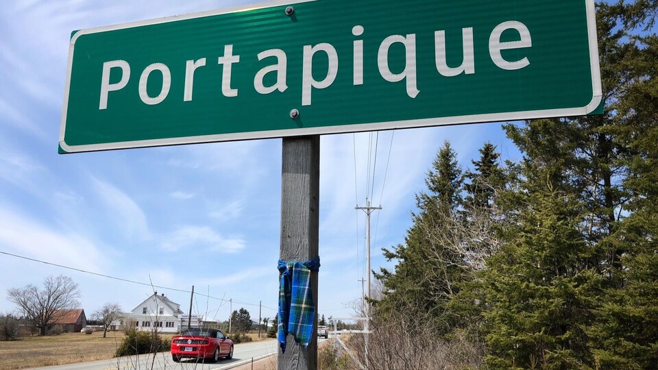 Tartan bleu et vert noué au poteau soutenant le panneau qui annonce l'entrée à Portapique, en bordure d'une route rurale.