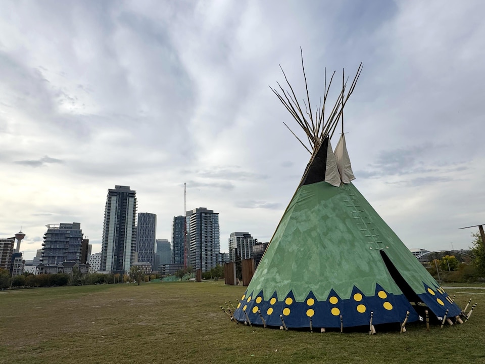 Un tipi vert et bleu, orné de motifs jaunes, se dresse sur une pelouse, avec des tours en arrière-plan sous un ciel nuageux.