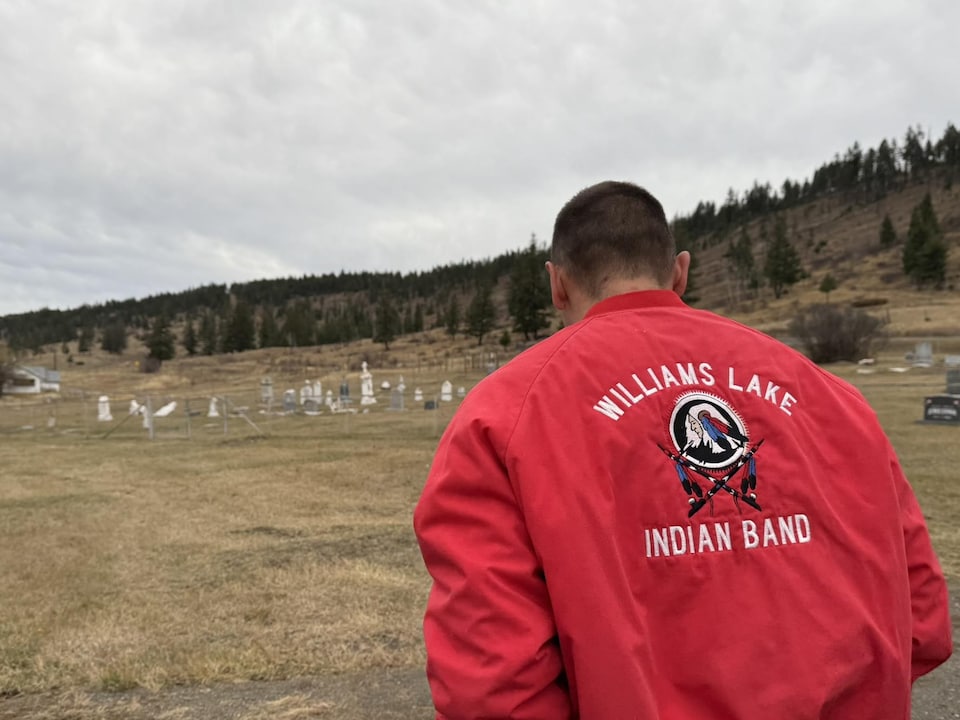 Un homme debout devant un cimetière est vêtu d'un blouson qui porte la mention Williams Lake Indian Band.