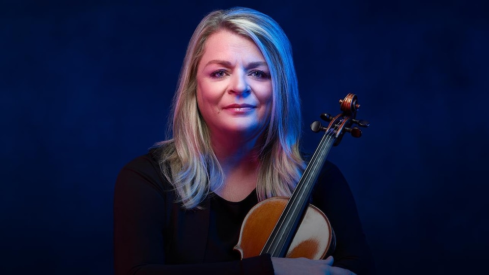 Une femme blonde posant de face, tenant un violon dans ses bras croisés
