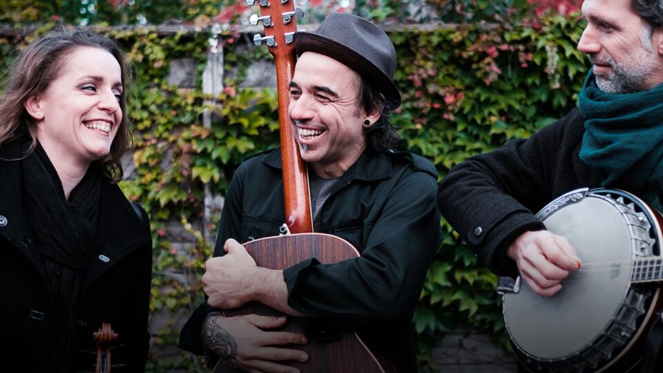 Les trois membres de la formation Bon Débarras (Marie-Pierre Lecault, Dominic Desrochers et Jean-François Dumas), à l'extérieur, tout sourire. Dominic, au centre, enlace sa guitare, alors que Jean-François, à droite, joue du banjo.