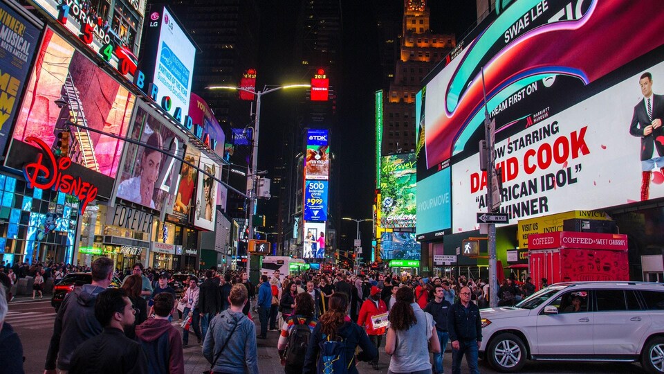 Nuit. Des centaines de passants marchent sur une rue bordée de nombreuses enseignes illuminées annonçant des spectacles à New York.