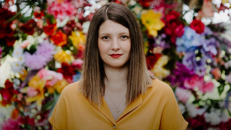 Chloé Breault pour "Plage des morons", son premier album complet (2020) : elle fixe l'objectif; cheveux droits; chemise jaune; elle se tient devant un mur de fleurs colorées.