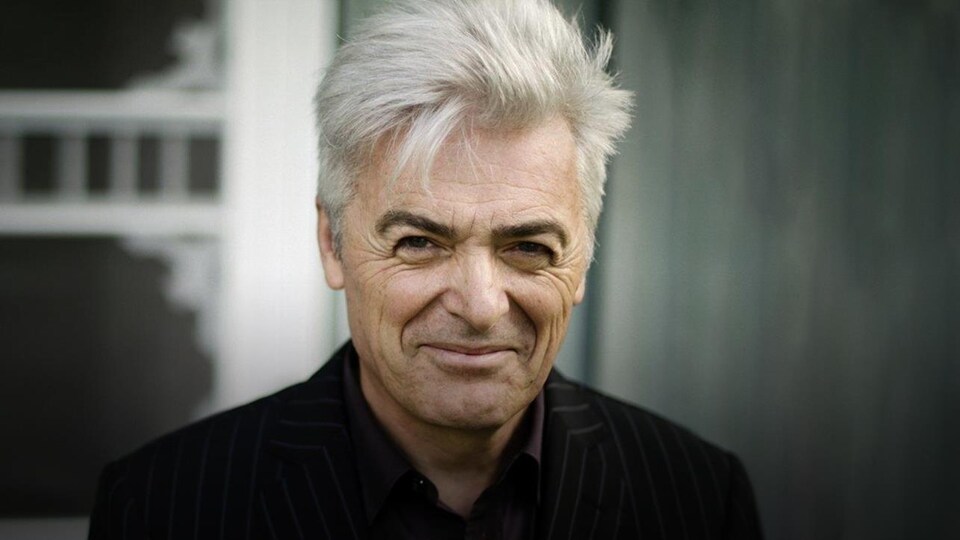 Daniel Lavoie souriant devant un arrière-plan gris et blanc très neutre.