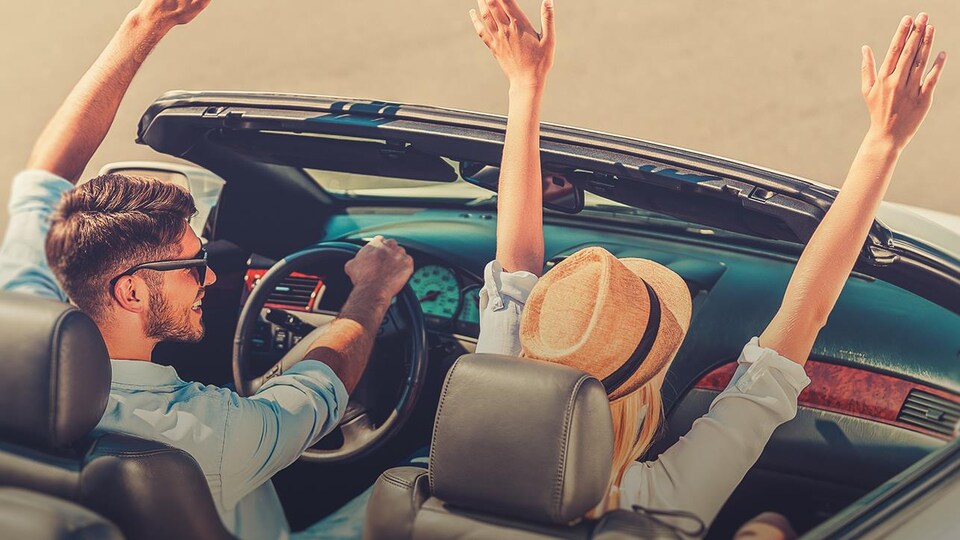 Un couple dans une voiture décapotable lèvent les bras.