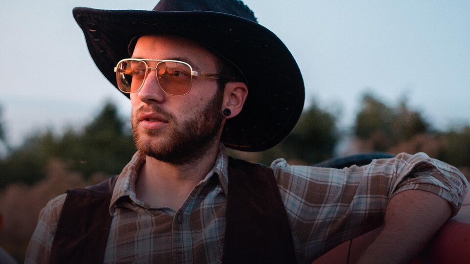 Menoncle Jason, lunettes de soleil, chapeau de cowboy et chemise à carreaux, le bras gauche appuyé sur une camionnette rouge.