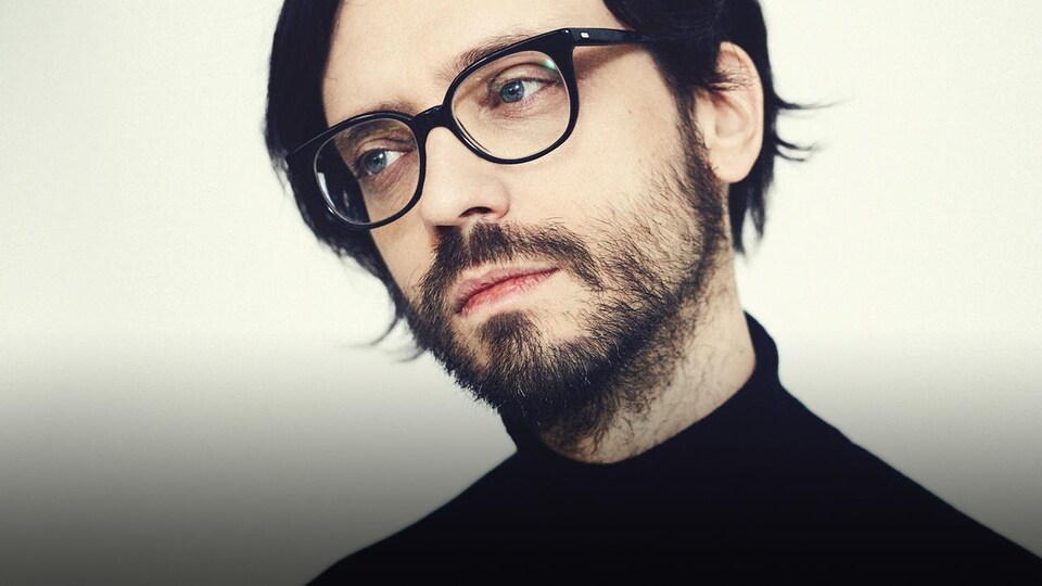 L'homme porte un col roulé noir, une barbe et des lunettes noires.