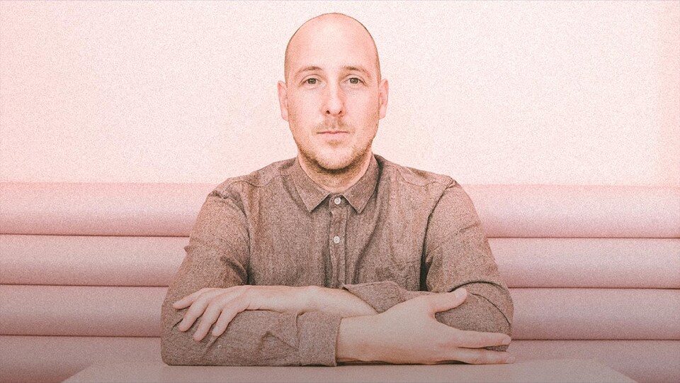 Le musicien Alex McMahon est attablé, assis sur une banquette rose, et regarde en face, les bras croisés. Il porte une chemise beige et a les cheveux rasés.