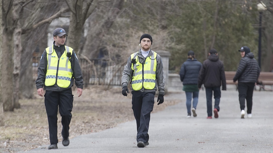 Deux cadets de la police marchent dans un parc, observés au loin par trois jeunes garçons.