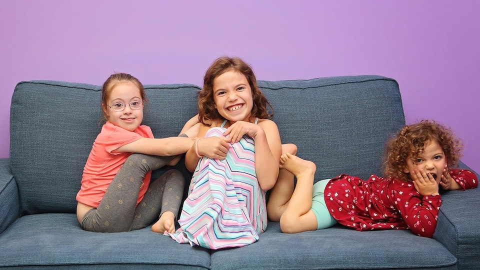 Trois jeunes filles assises sur une causeuse grise regardent la caméra en souriant.