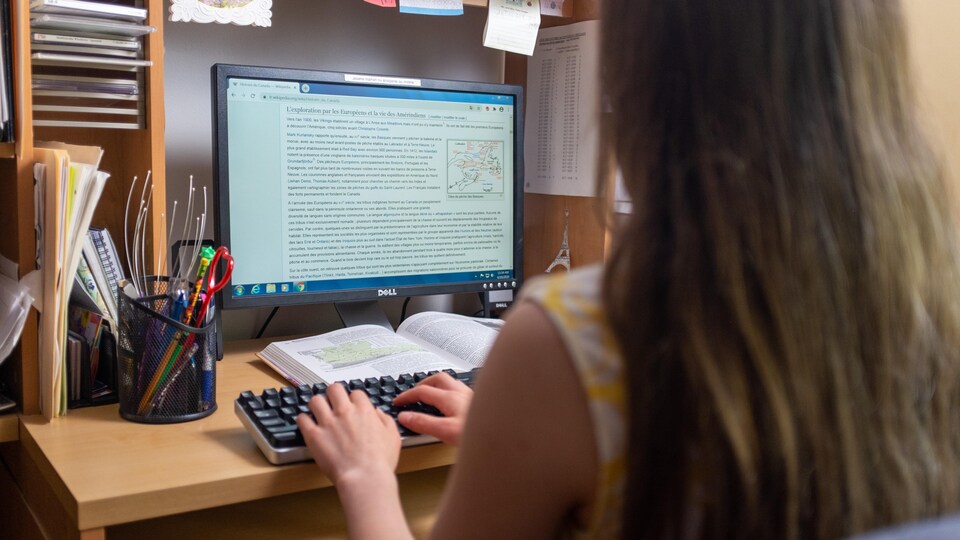 L'adolescente est devant un ordinateur sur un bureau avec un cahier d'apprentissage et du matériel scolaire. 