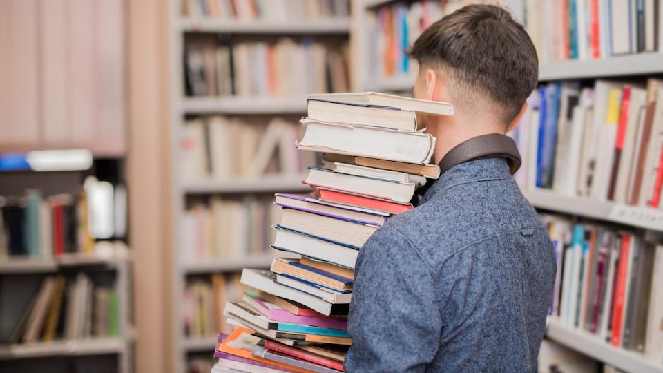 Un jeune homme vu de dos, portant de nombreux livres en équilibre, dans une bibliothèque.