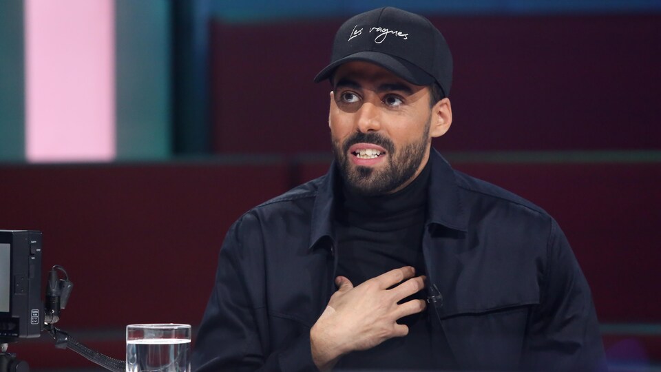 Adib Alkhalidey est à la table des invités. Il porte une casquette, une veste et un chandail noirs.