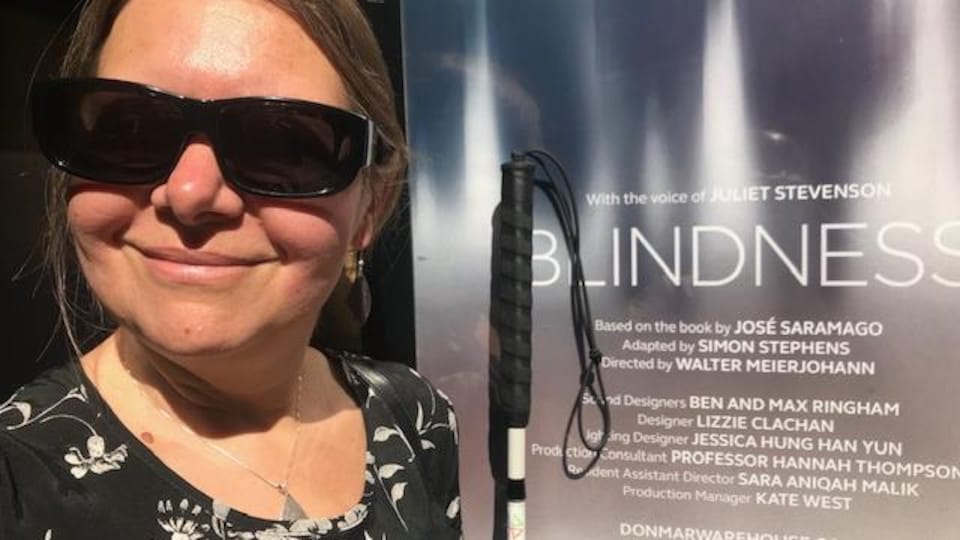 Hannah Thompson posée devant l'affiche de BLINDNESS, qui énumère les membres de la distribution et de l'équipe de production, y compris la photographiée. Elle est souriante, lunettes fumées aux yeux et canne blanche à la main.