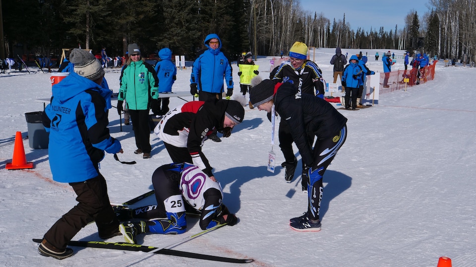 Des jeunes athlètes et spectateurs sont à l'extérieur sur une surface enneigée à l'occasion des Jeux d'hiver de l'Arctique. Un compétiteur qui vient de terminer sa compétition est sur le sol et des personnes sont près de lui pour l'encourager.