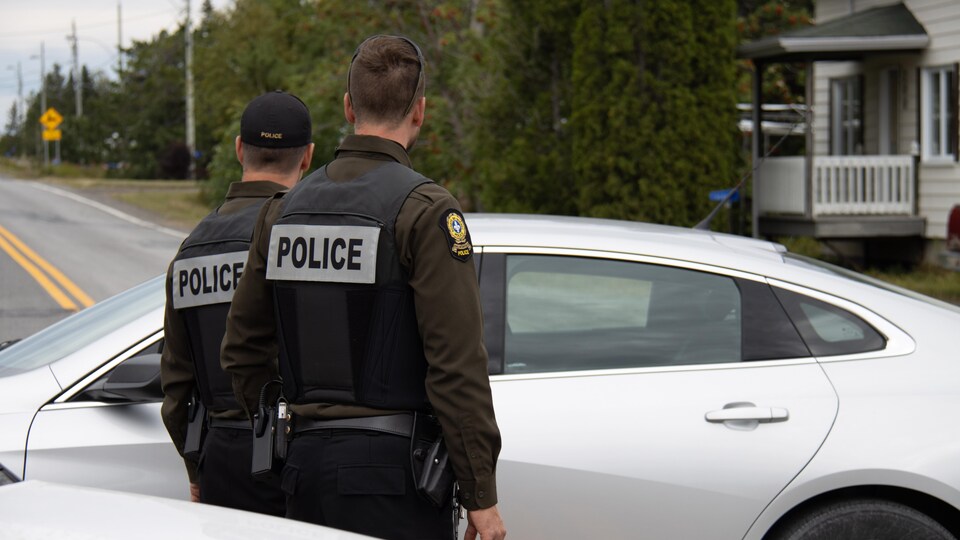 Two policemen, from behind, in front of a car on a country road.