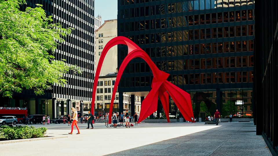 Une place publique qui a en son centre une structure de metal, un stabile, peinte en rouge.