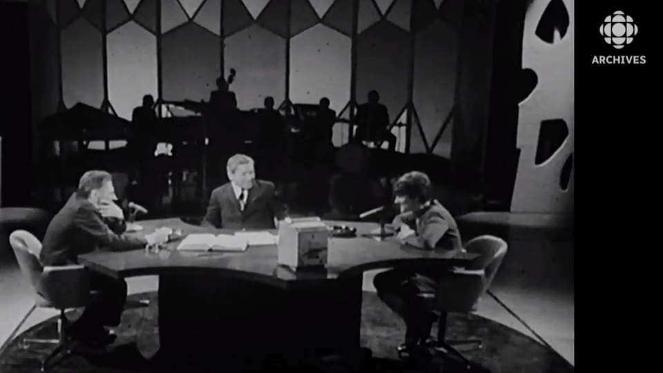Deux hommes et une femme discutent, assis à une table ronde, sur un plateau de télévision.