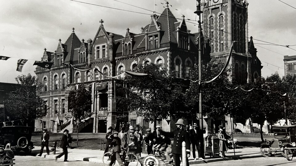 Photo en noir et blanc de l’ancien hôtel du ville de Regina. Il y a des pietons, des gens à vélo, des voitures.