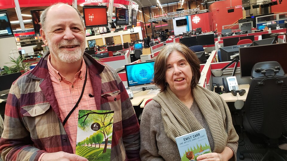 Un quinquagénaire souriant tenant un catalogue et une sexagénaire tenant son livre debout dans une salle de nouvelles.