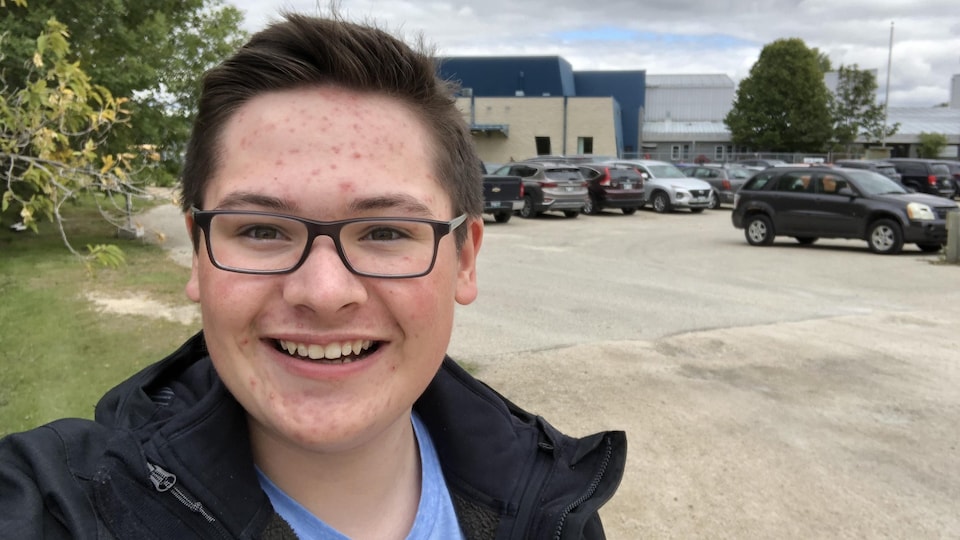 André Boisjoli se prend un égoportrait devant une école où sont aussi statitionnés des véhicules