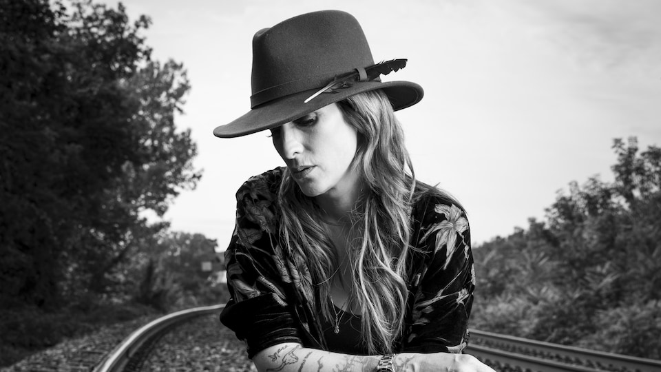 Photo en noir et blanc d'une femme portant un Stetson et assise sur un étui de guitare au milieu de rails de chemin de fer.