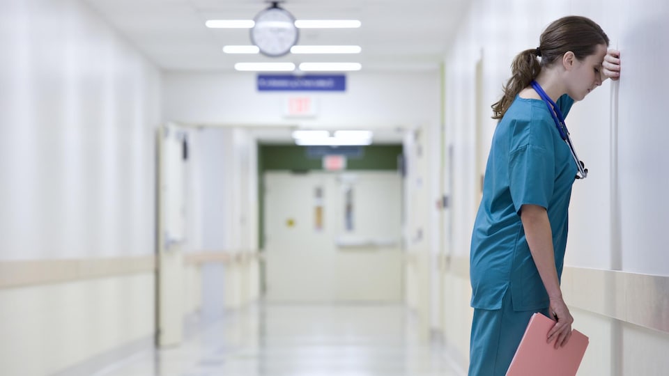 Une professionnelle de la santé est appuyée contre un mur dans un couloir d'hôpital.