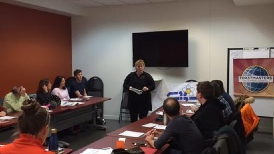 Un groupe de personnes assises à des bureaux écoutent une femme debout au fond de la pièce.