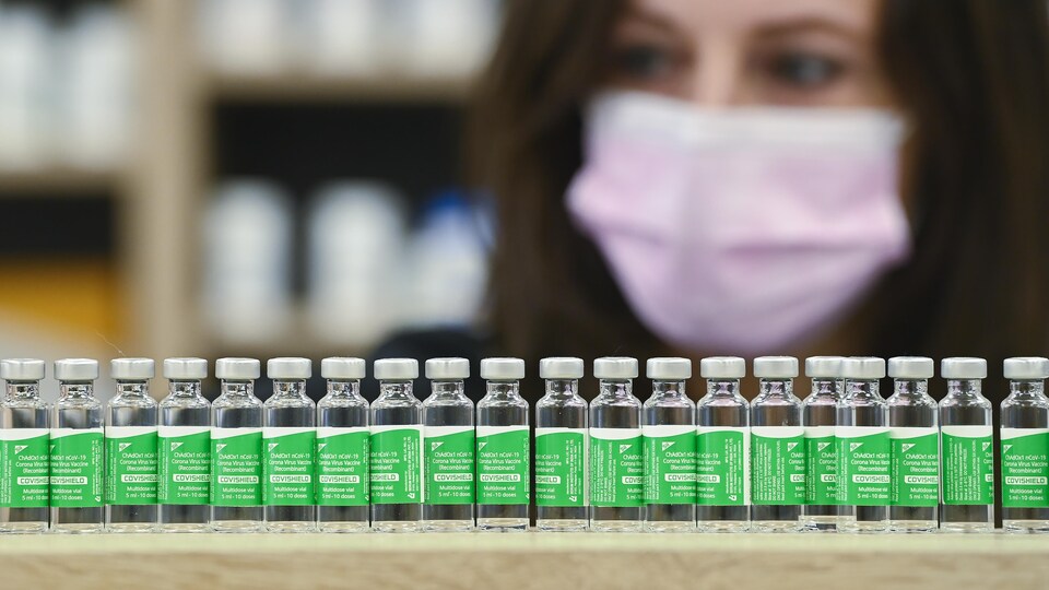 Gros plan du visage d'une femme qui porte un masque médical, avec en avant-plan une rangée de fioles de vaccin AstraZeneca.