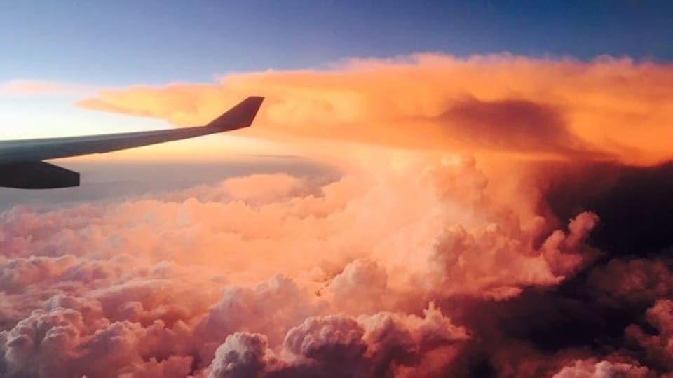Photo prise à bord d'un avion en vol montrant le reflet orangé du soleil sur les nuages et le bout d'une aile de l'avion.