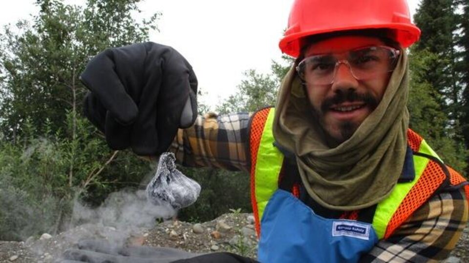 Un chercheur est dans la foret avec un casque route et tient avec des gros gants un objet qui fume.