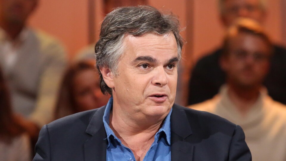 L'homme parle. Il porte une chemise bleue et un veston noir.